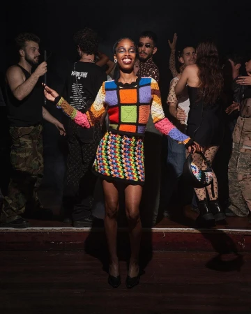 An adult woman in a colorful crochet sweater and skirt poses on a stage in front of a crowd of people in a dimly lit room.
