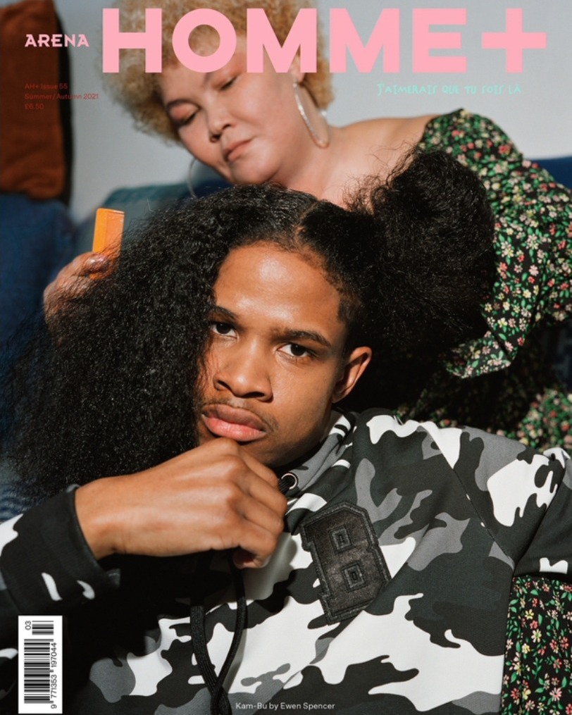 Magazine cover featuring a young man in a camo hoodie being styled by an older woman with an orange comb indoors.