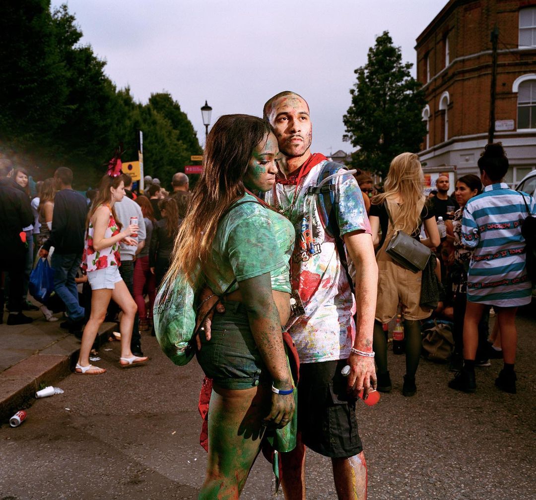A couple, an adult man and woman, covered in paint embrace in an outdoor street scene with a crowd and buildings in the background.