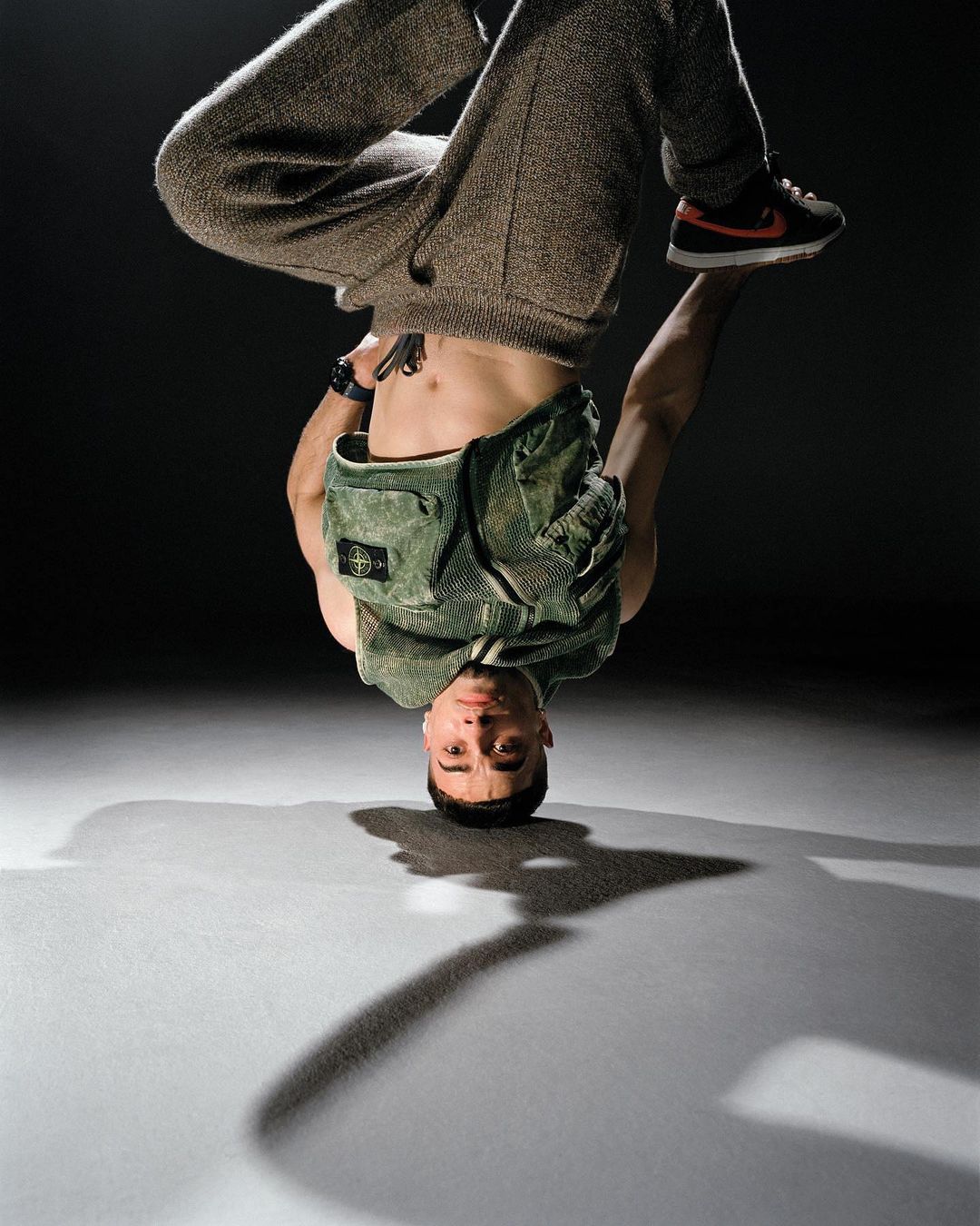 Male dancer in a headstand wearing an olive-green vest and gray pants. Feet in the air. The studio has a dark backdrop.
