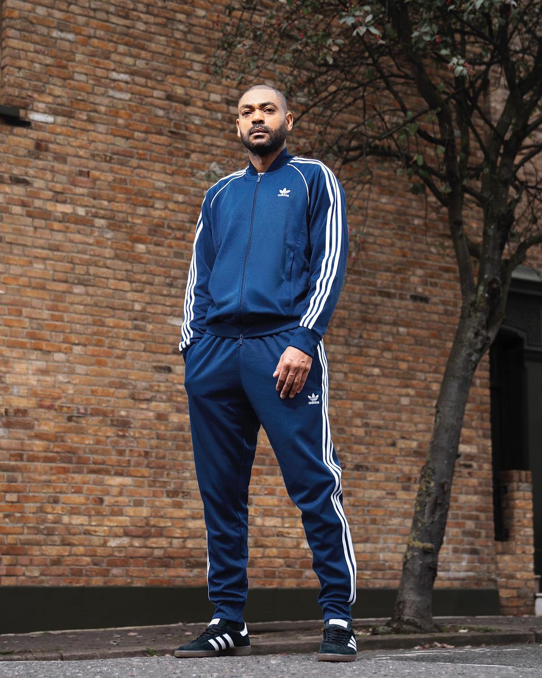 A man in a blue Adidas tracksuit stands in front of a brick wall with a tree, posing for a fashion or lifestyle portrait.