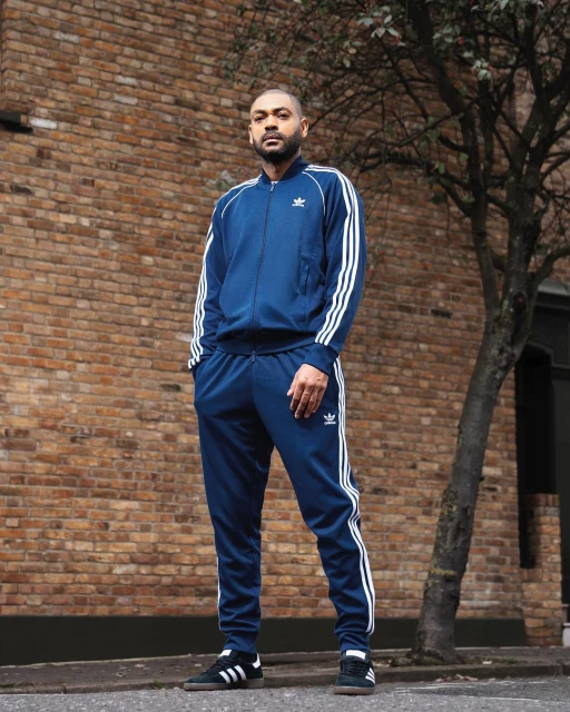 A man in a blue Adidas tracksuit stands in front of a brick wall with a tree, posing for a fashion or lifestyle portrait.