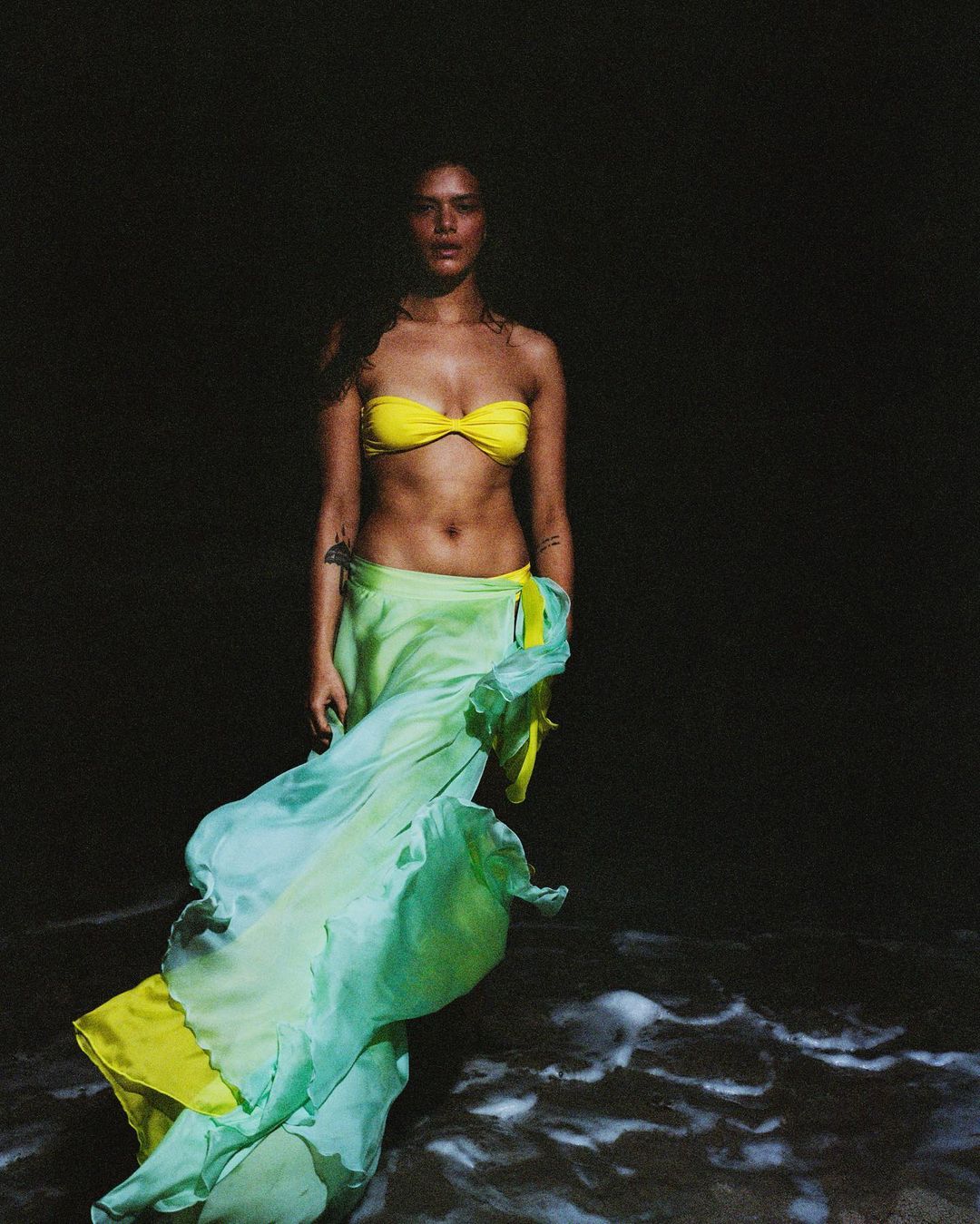 Adult woman in yellow bikini top and green skirt stands on dark sandy beach with foamy waves; dark background, medium shot.