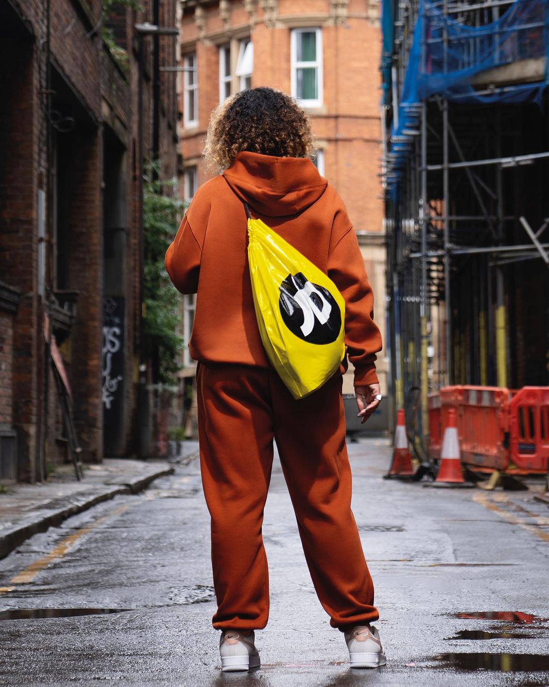Adult with curly hair walks down wet city alley with a bright yellow shoulder bag, wearing a matching rust-colored sweatsuit.