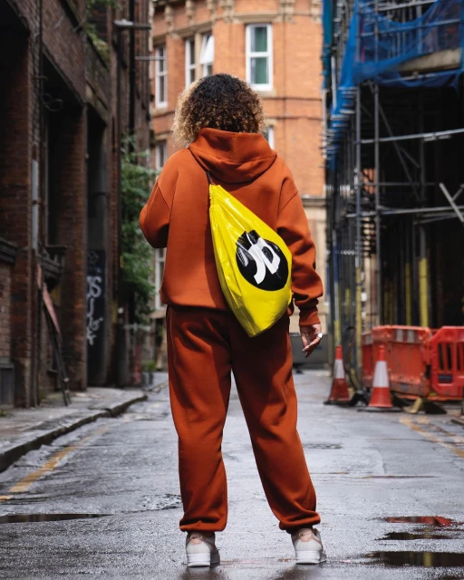 Adult with curly hair walks down wet city alley with a bright yellow shoulder bag, wearing a matching rust-colored sweatsuit.