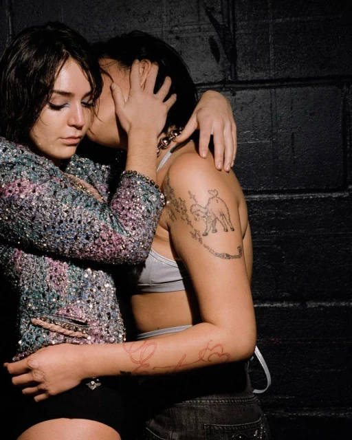 Two young women embracing in front of a textured dark wall. One has arm tattoos, the other wears a sparkling jacket.