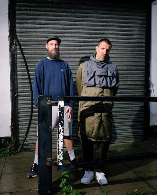 Two adult men pose outdoors in casual streetwear in front of a corrugated metal wall, alongside a black metal railing and urban sidewalk.