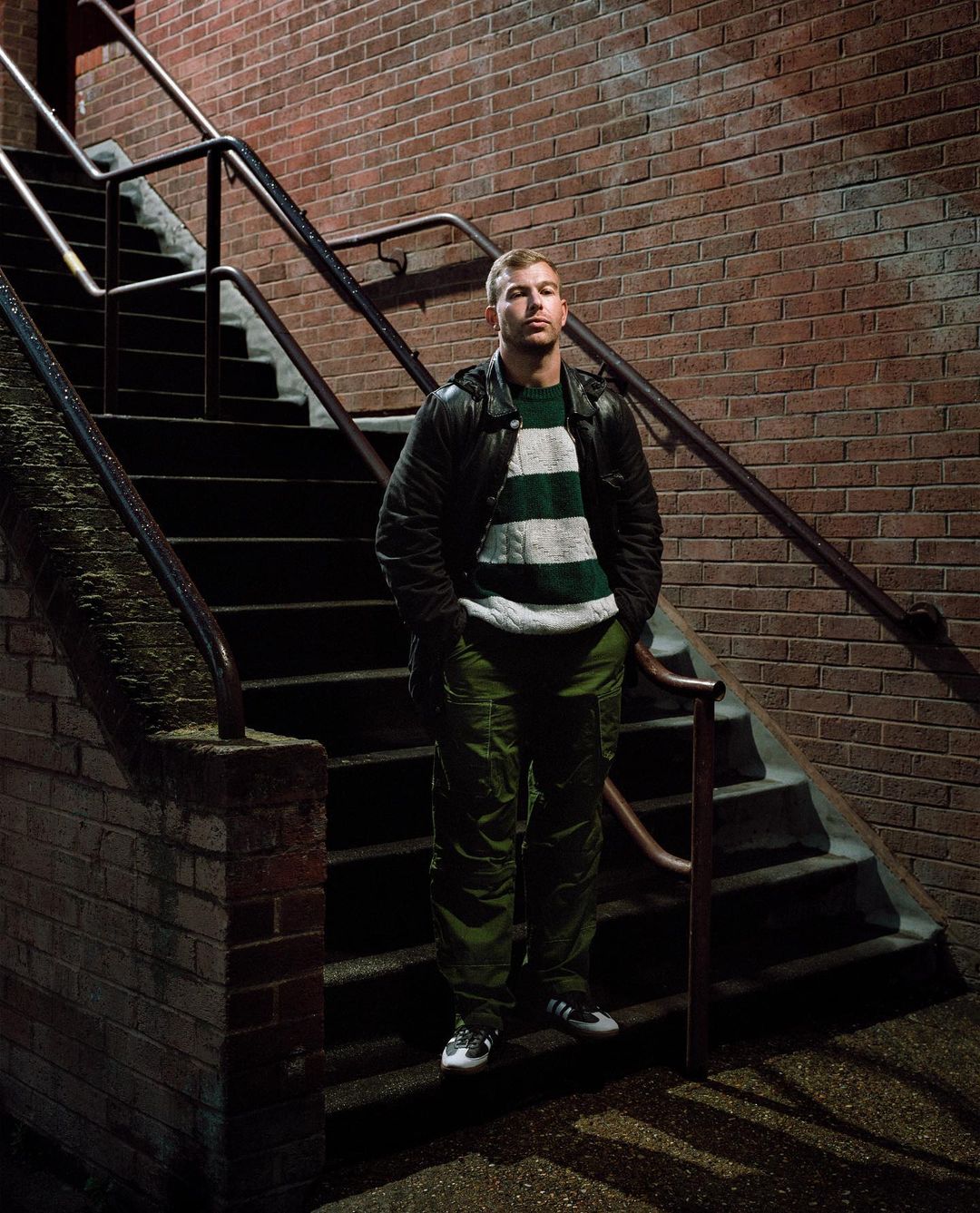Man stands on steps in green pants and striped sweater near brick wall. Urban scene with dramatic lighting. Fashion portrait.