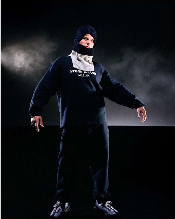 Full-length studio shot of an adult male wearing a dark sweatshirt, sweatpants, sneakers, and a knit balaclava against a dark background.