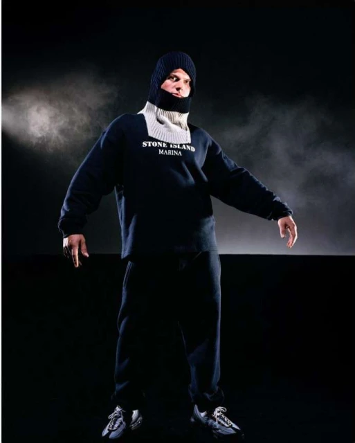 Full-length studio shot of an adult male wearing a dark sweatshirt, sweatpants, sneakers, and a knit balaclava against a dark background.