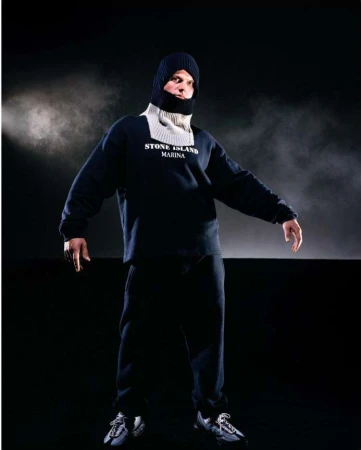 Full-length studio shot of an adult male wearing a dark sweatshirt, sweatpants, sneakers, and a knit balaclava against a dark background.