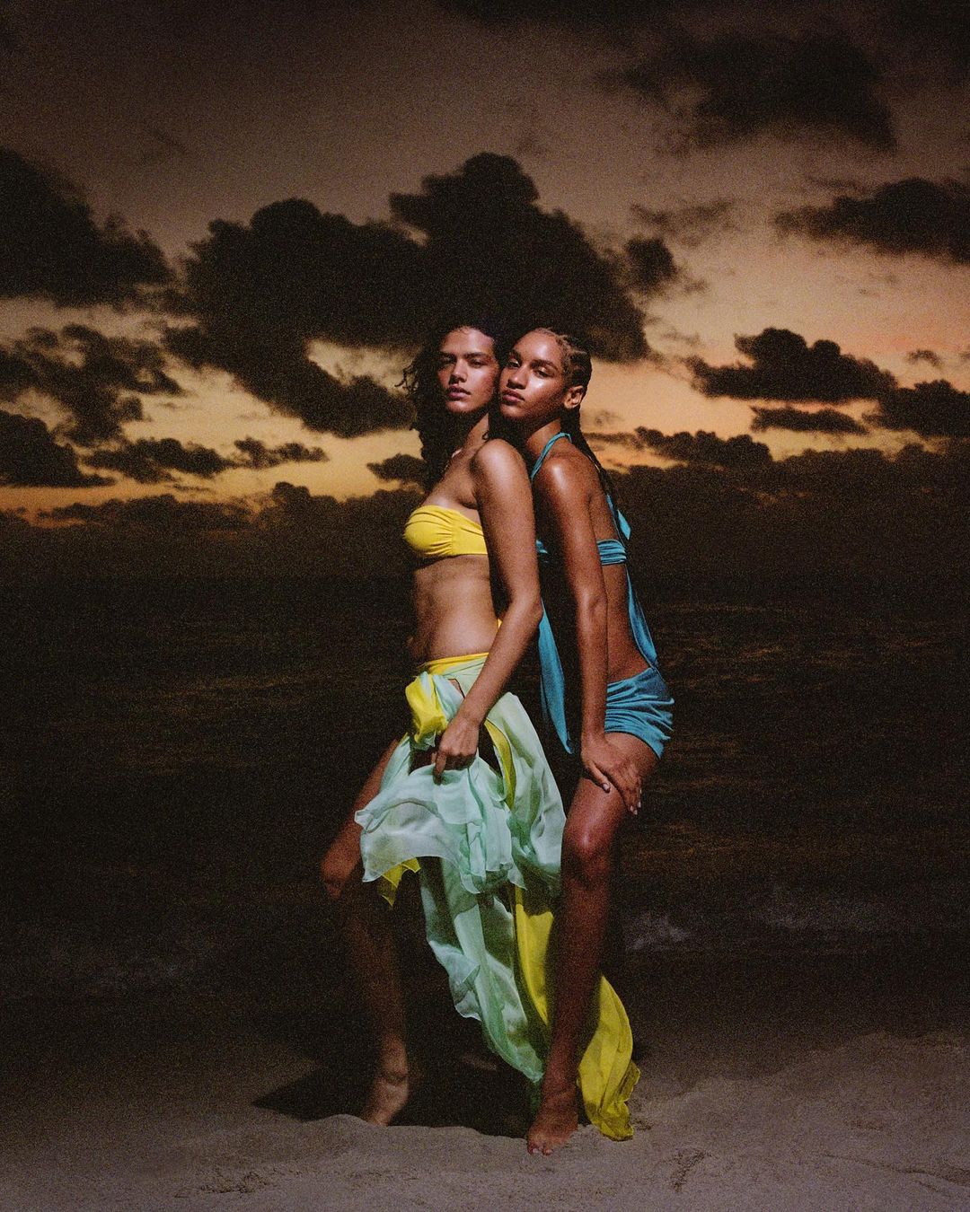 Two women pose in bikinis and draped fabrics on a beach against a sunset sky. Fashion shot with sand, clouds, and ocean backdrop.