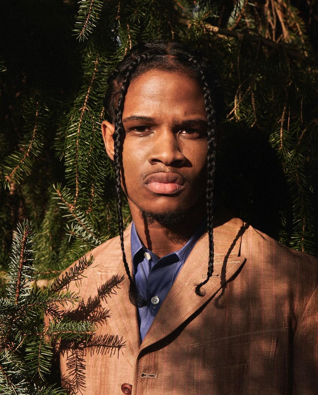 Portrait of a Black male with braided hair wearing a blazer and button-down shirt, standing in a natural setting with evergreen trees.