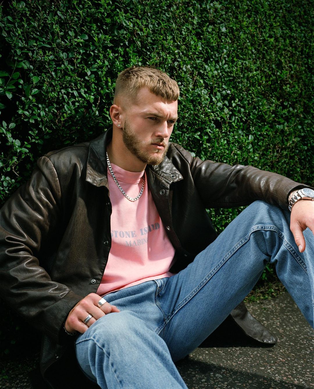 Man in leather jacket and jeans sits against a green hedge, wearing a pink t-shirt and silver chain. He is looking off to the side.