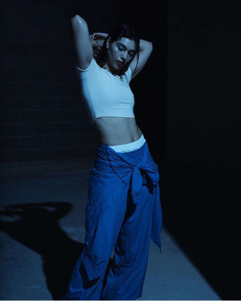 Brunette woman poses indoors wearing a white crop top and tied blue pants with hands behind her head in a dimly lit setting.