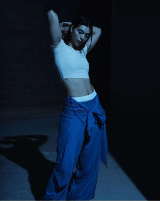 Brunette woman poses indoors wearing a white crop top and tied blue pants with hands behind her head in a dimly lit setting.