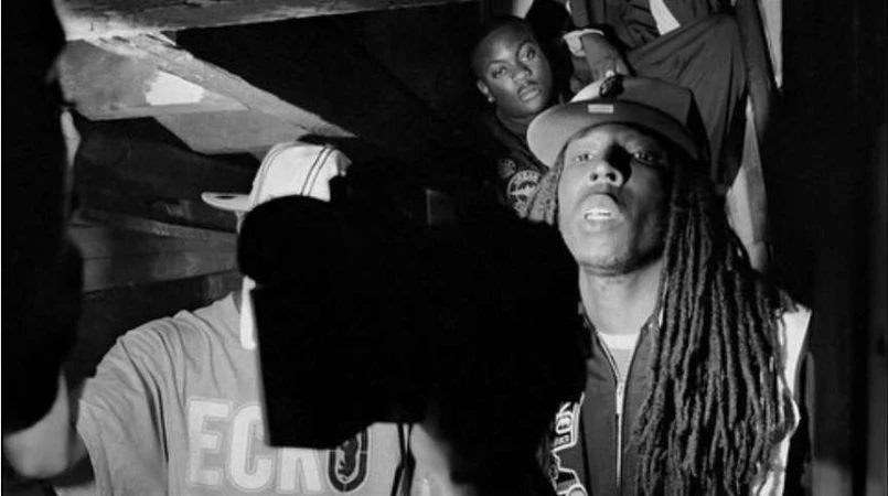 Monochrome image of three African American men wearing streetwear-style clothing, indoors under a low ceiling, with dark shadows.