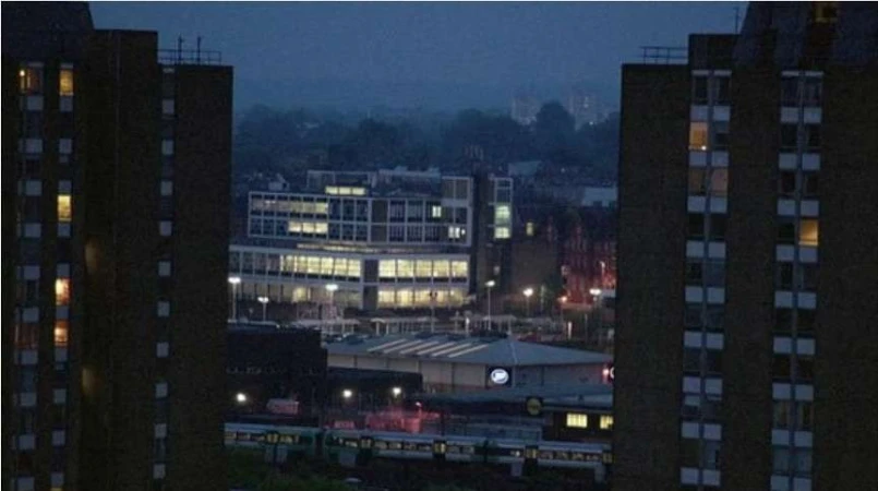 Cityscape at night with illuminated buildings, viewed between two tall apartment buildings, creating a framed urban scene in the darkness.