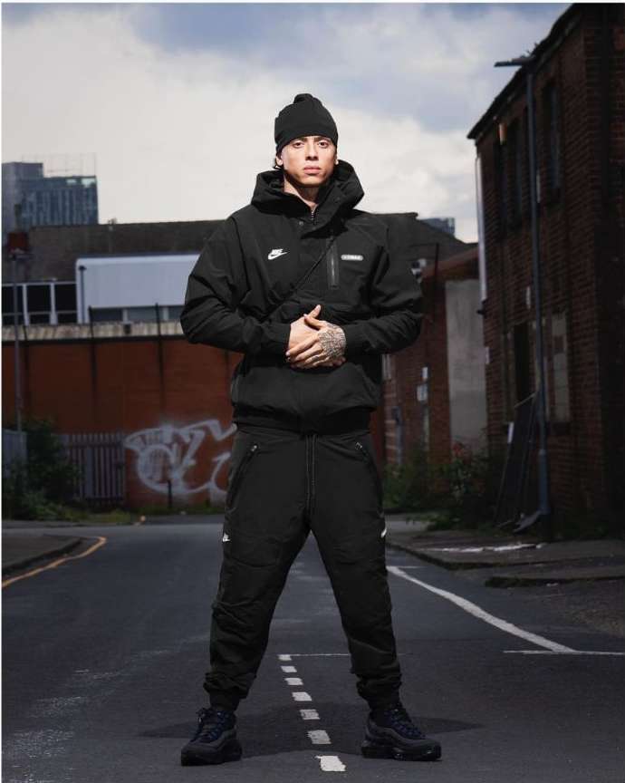 Man in black sportswear stands on a city street. He wears a jacket, beanie, pants, and sneakers. Urban environment with buildings behind.