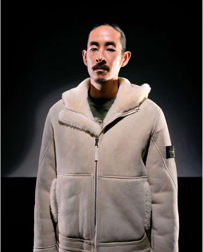 Portrait of an adult man with dark hair and a mustache wearing a light beige shearling bomber jacket against a dark background.