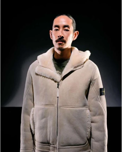 Portrait of an adult man with dark hair and a mustache wearing a light beige shearling bomber jacket against a dark background.