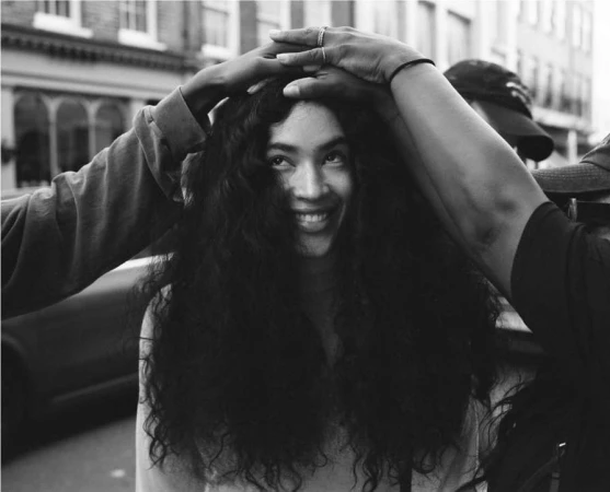 Black and white portrait of a smiling young woman with long curly hair framed by hands touching her head.