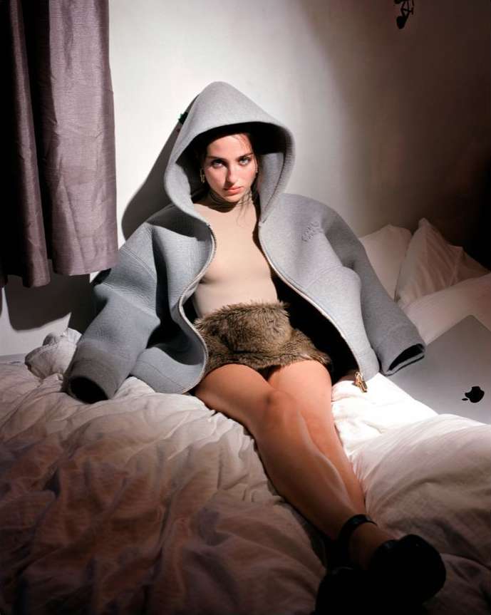 Woman poses on a bed in an oversized light gray hooded jacket with fur skirt, looking at the camera in an indoor studio shot.