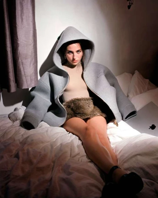 Woman poses on a bed in an oversized light gray hooded jacket with fur skirt, looking at the camera in an indoor studio shot.