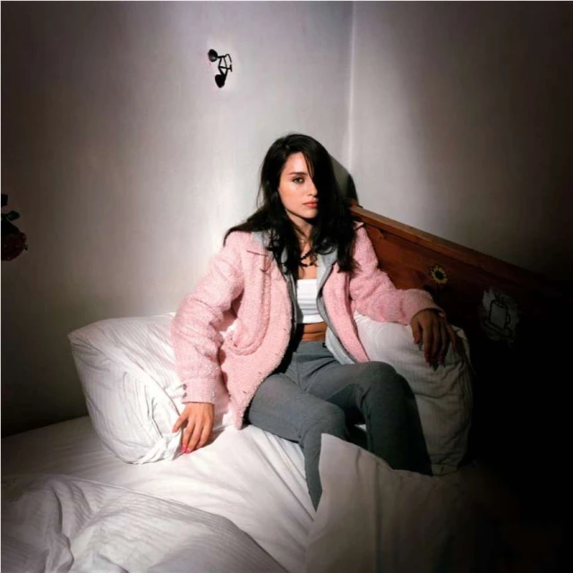 A woman with long dark hair sits on a bed with white linens wearing a pink jacket and grey pants in an indoor setting with dark shadows.