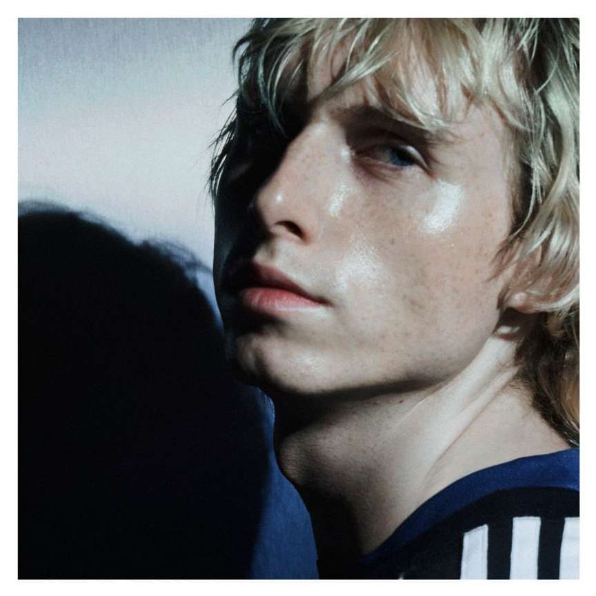 Portrait of a young adult with blond hair wearing a blue and white striped athletic top, posing in a brightly lit studio setting.