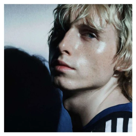 Portrait of a young adult with blond hair wearing a blue and white striped athletic top, posing in a brightly lit studio setting.