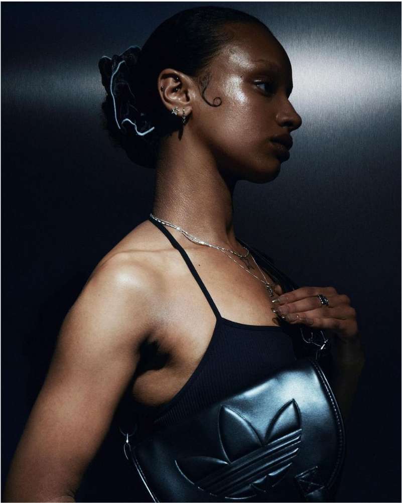 Profile portrait of an adult woman with sleek hair, wearing a black halter top with an Adidas logo, against a dark gray background.
