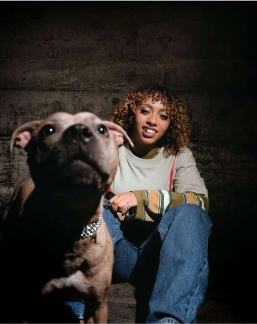 Young woman with blonde curly hair poses while seated next to her tan pitbull dog indoors in a dimly lit room with brick wall.