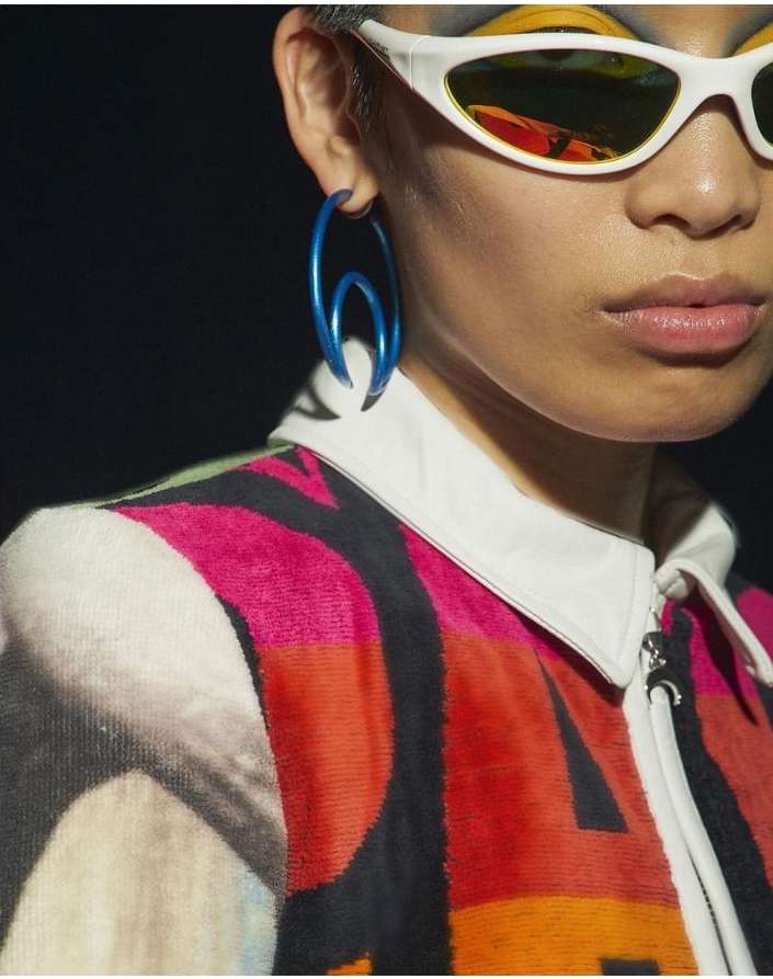 Close-up shot of a model showcasing an oversized, unique blue hoop earring, wearing sunglasses and a vividly patterned, zippered jacket.