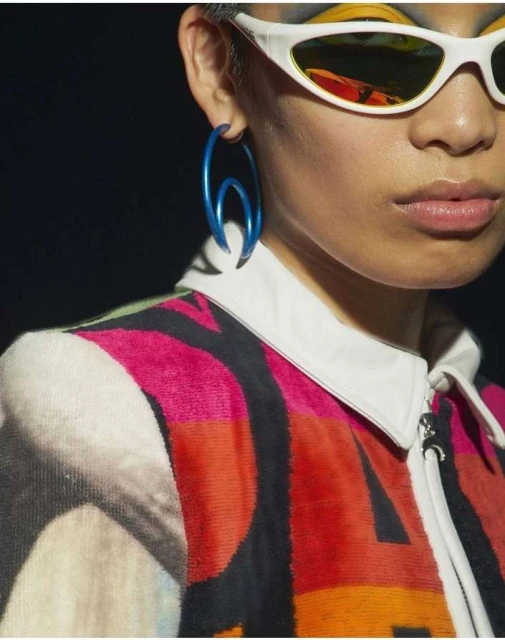 Close-up shot of a model showcasing an oversized, unique blue hoop earring, wearing sunglasses and a vividly patterned, zippered jacket.