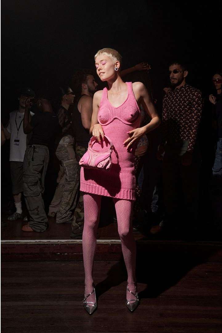 Woman in a pink knit dress and heels posing with a purse in a crowded indoor setting with a dark ambiance and warm lighting.