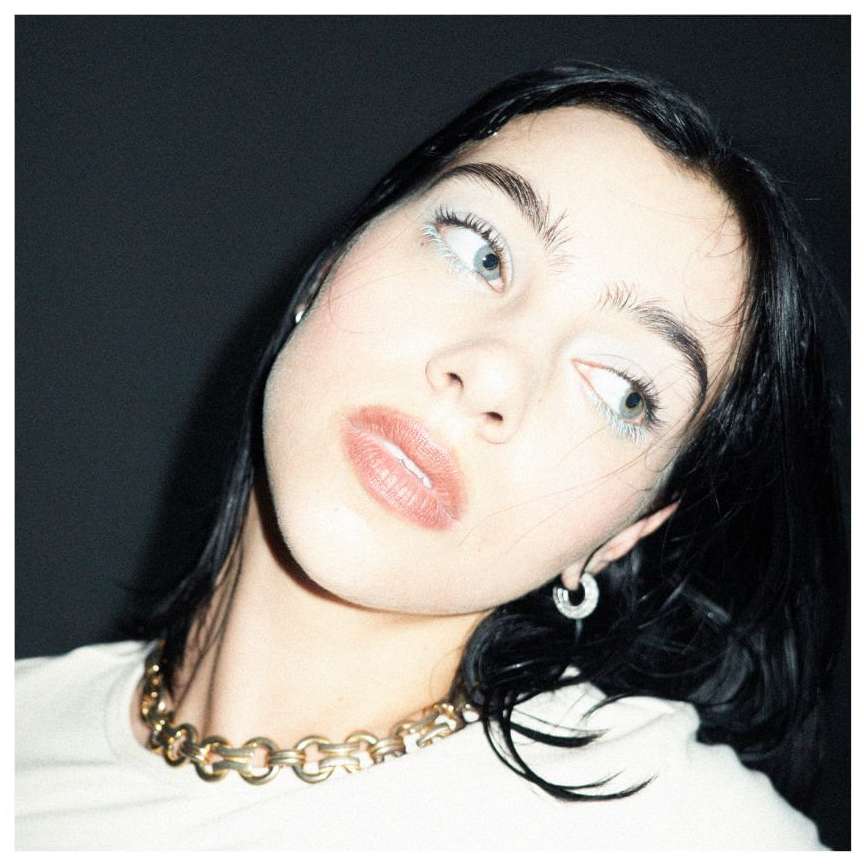 Close-up shot of a young woman with fair skin, dark hair, wearing a gold chain necklace against a dark background, looking up and away.