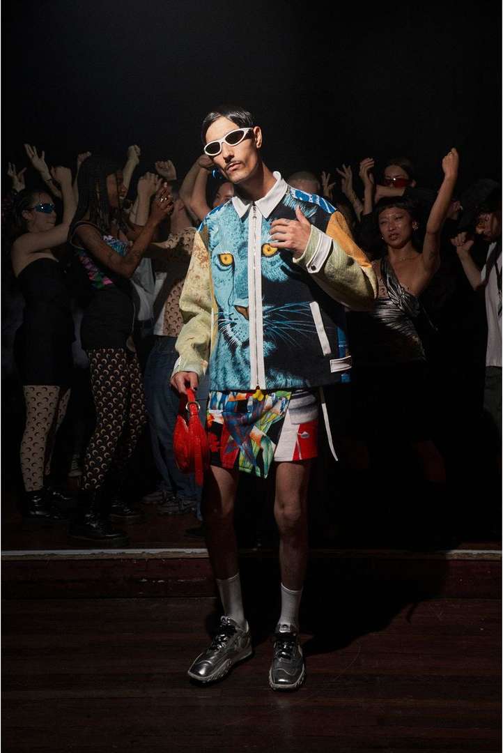 Stylish adult male poses in a patterned jacket and shorts, with a crowd of cheering people in the dark background at a club or event.
