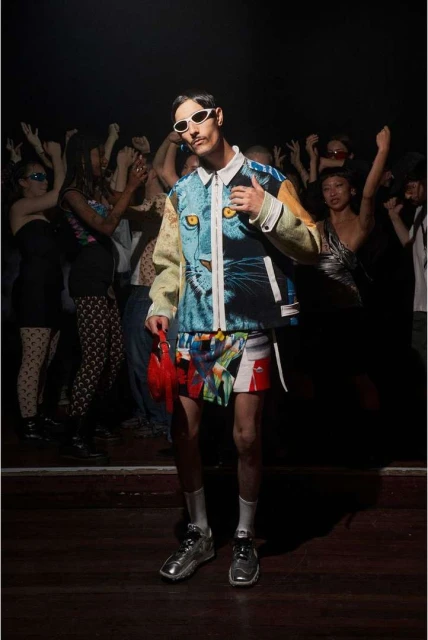 Stylish adult male poses in a patterned jacket and shorts, with a crowd of cheering people in the dark background at a club or event.