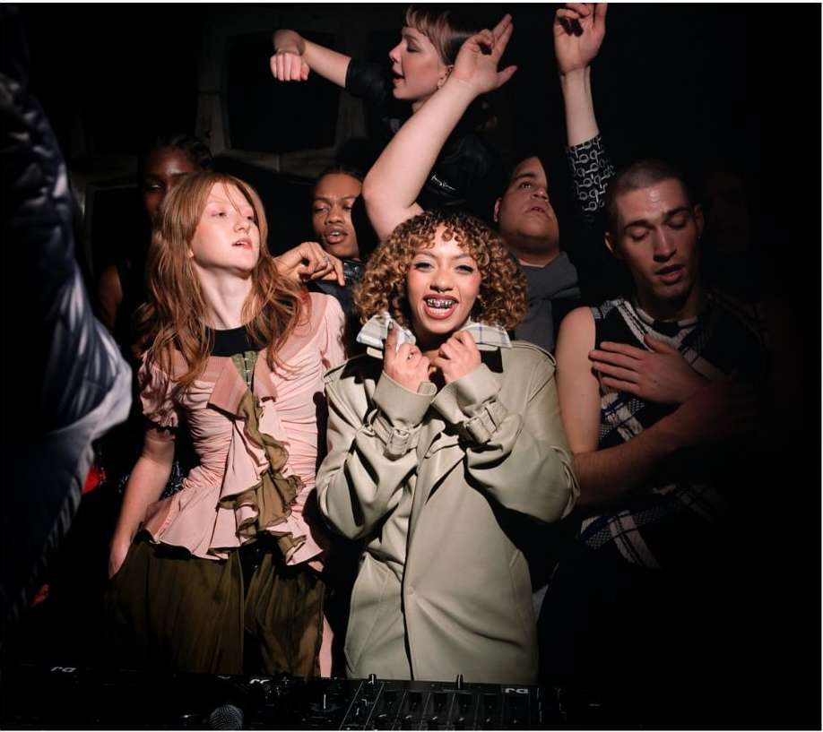 Group of people in a dimly lit indoor club setting, wearing fashionable clothing. The image is a candid shot with warm color grading.