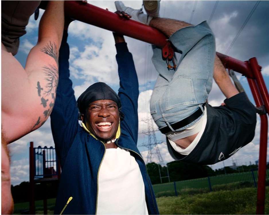 Three men are on a playground jungle gym, one hanging upside down, another smiling while holding onto the bar with a tattoo on their arm.