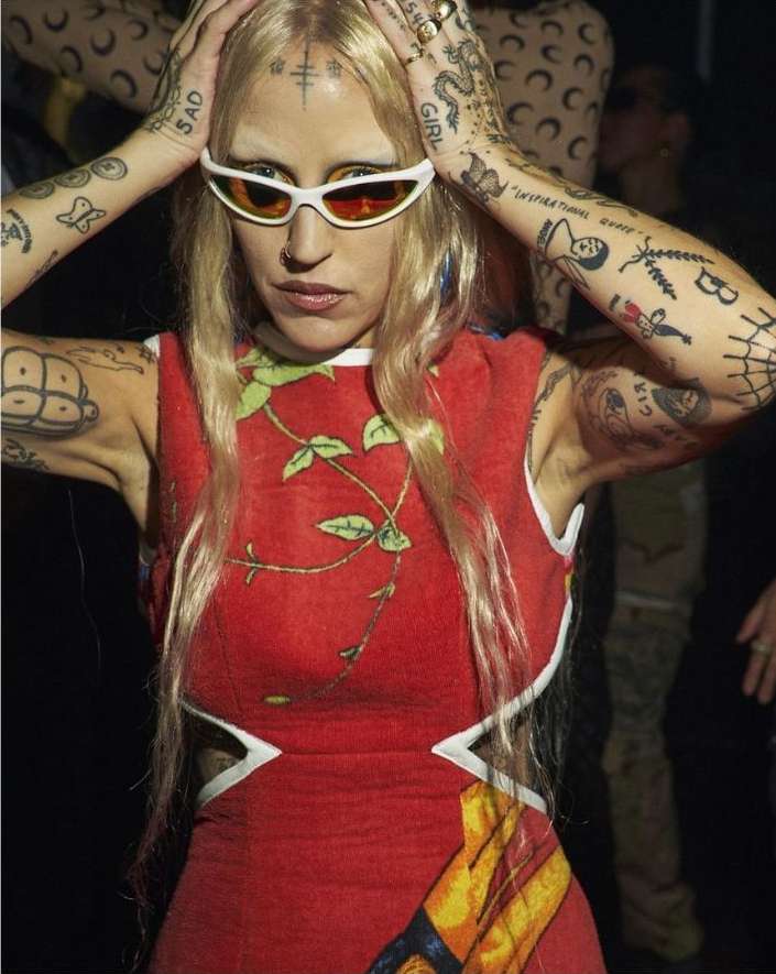 Blonde woman with arm tattoos wears a floral red dress and sunglasses indoors, hands above her head in a medium shot.