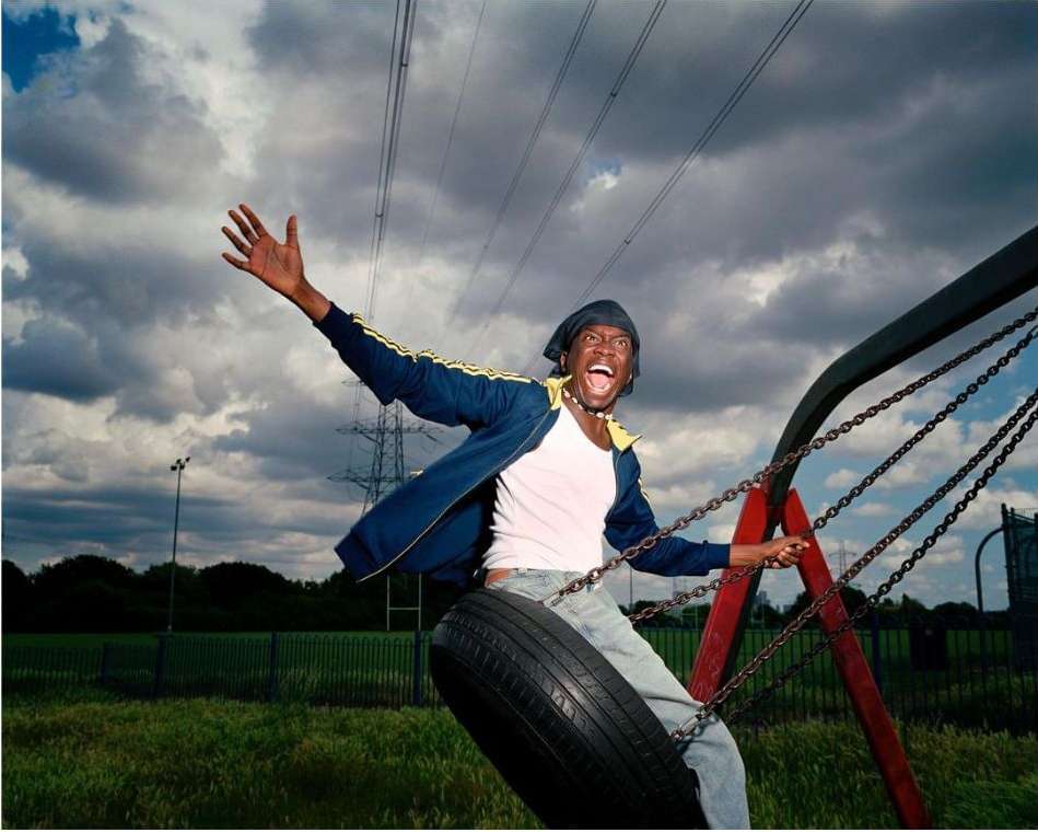 Enthusiastic African American man on a tire swing, arms raised with an open mouth, wearing a blue jacket and jeans, grassy field and cloudy sky backdrop.