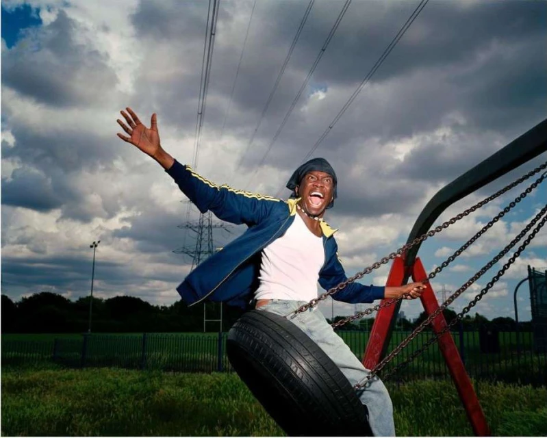 Enthusiastic African American man on a tire swing, arms raised with an open mouth, wearing a blue jacket and jeans, grassy field and cloudy sky backdrop.