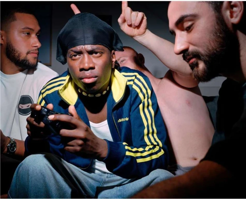 A group of four men hanging out indoors. One man plays a video game with a dark controller, with other friends nearby.