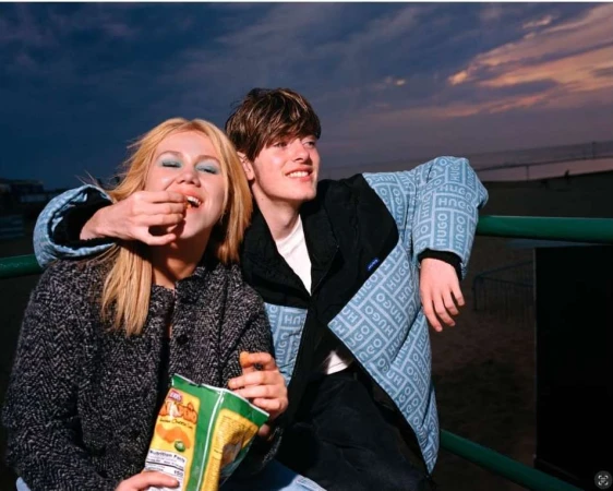 Young couple snacking outdoors at dusk; a young woman is fed by a young man wearing a Hugo jacket, with ocean and twilight background.
