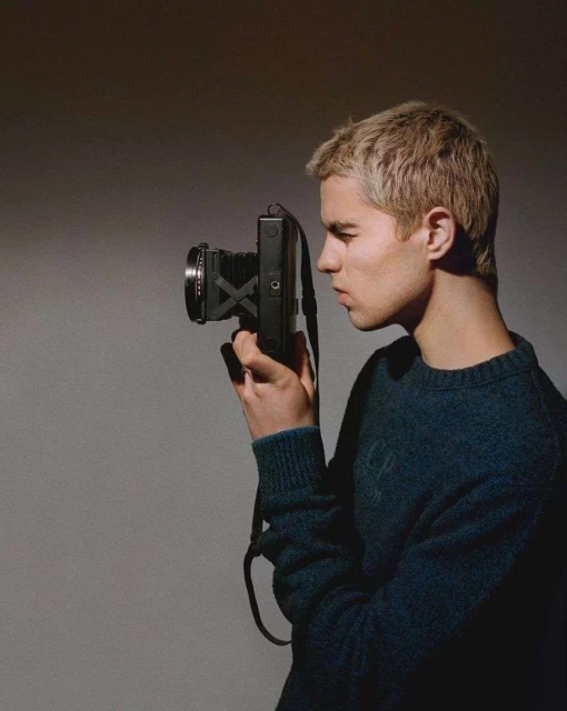 Young adult male with blond hair looking through the viewfinder of a vintage camera in a neutral-toned studio setting.