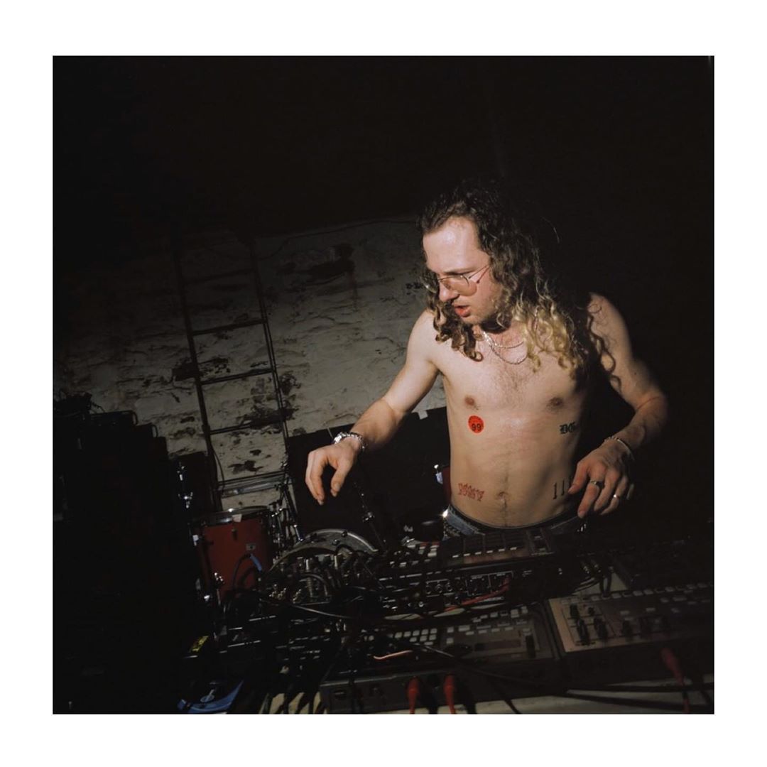A shirtless man with long hair wearing glasses adjusting audio equipment in a dark room with exposed brick and dim lighting.