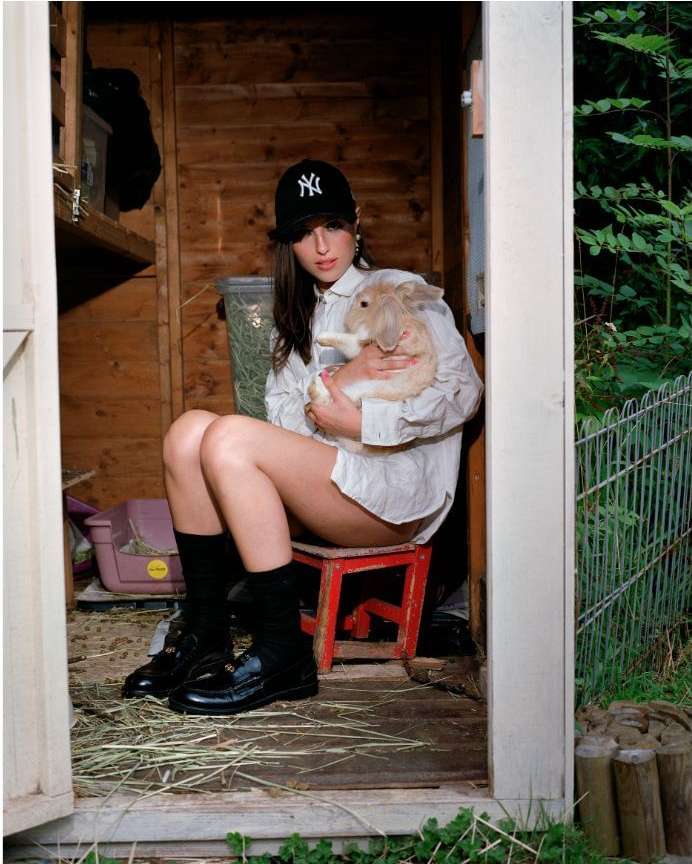 Young woman in baseball cap sits on a red stool, inside a wood shed, cradling a beige rabbit, surrounded by greenery in an outdoor setting.