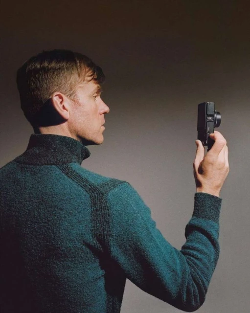 Adult male in a teal sweater holding a small camera, back to the viewer, looking left toward the camera, against a grey background.